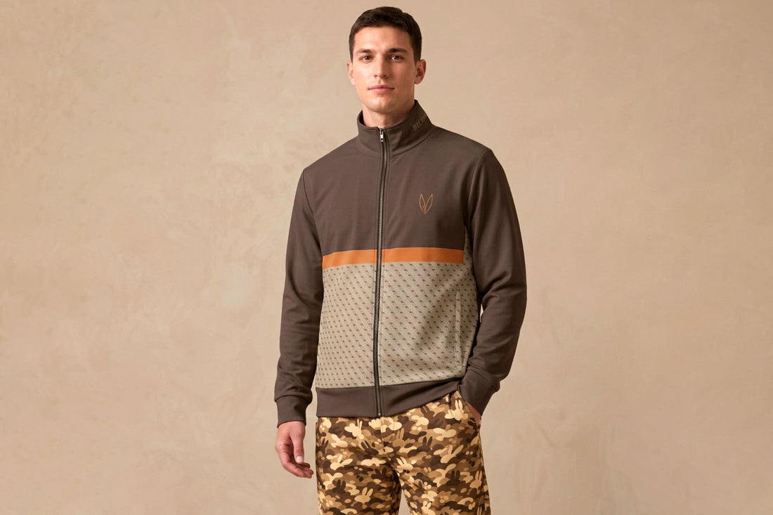 Male model in dark zip track jacket with amber detail and camouflage trousers against a warm neutral background — cold weather outfit editorial, Biliping