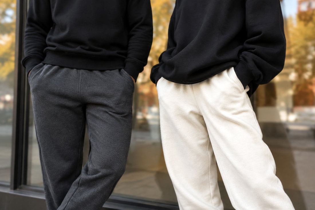 Two people in black sweatshirts and neutral joggers leaning against a glass storefront window with blurred autumn street in the background — October Dressing editorial, Biliping