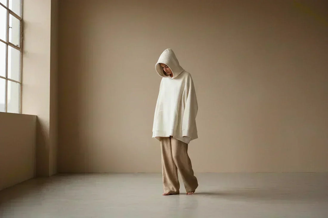 Person in oversized ivory hoodie and sand wide-leg pants standing quietly in a warm minimal room — fashion as personal transformation